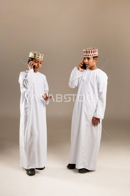 Using a modern and advanced technical device, entertainment and recreation in free time, portrait of two Omani Gulf Arab boys wearing a dishdasha and a kummah, one of them holding a mobile phone in his hand and making a phone call, full body, beige background.