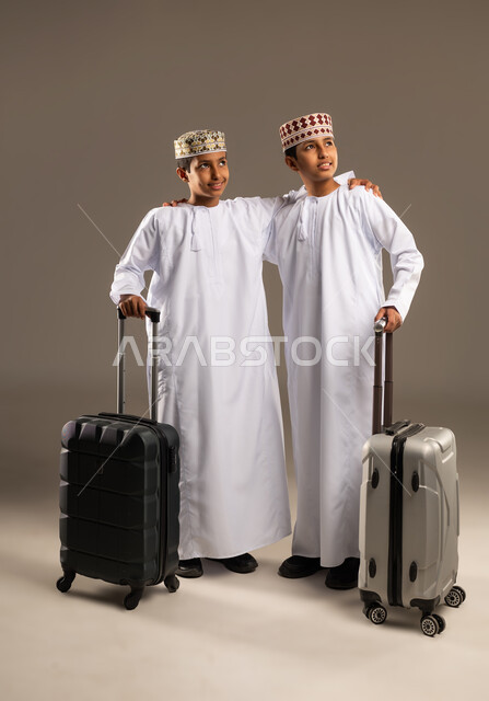 النظر الى شئ ما فى الاعلى ، قضاء رحلة سياحية ممتعة في إجازة الصيف، بورتريه لولدان توأم عربيان خليجيان عمانيان يرتديان الدشداشة والكمة يمسكان حقائب السفر ، صورة لكامل الجسم، خلفية بيج .
