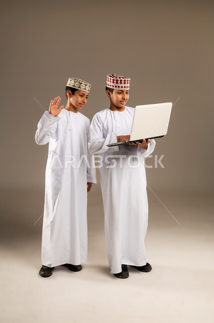 اجراء مكالمات الفيديو بإيماءات السرور والاندماج، الاستفادة من التقنيات الحديثة في تزويد الأجيال بالمعلومات المفيدة، بورتريه لولدان توأم عربيان خليجيان عمانيان يرتديان الدشداشة والكمة احدهما يقوم باستخدام جهاز الحاسوب المحمول، خلفية بيج .