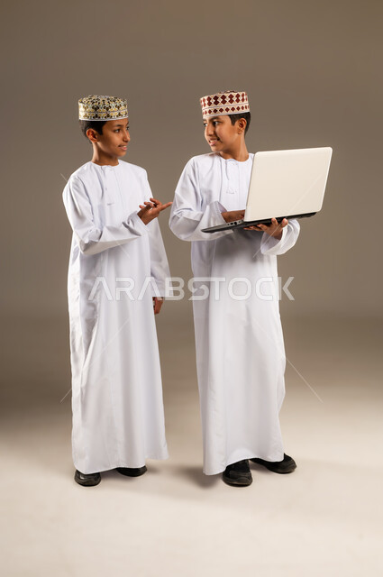 الاستفادة من التقنيات الحديثة في تزويد الأجيال بالمعلومات المفيدة، متابعه البرامج الترفيهيه المفضلة بإيماءات السرور والاندماج، بورتريه لولدان توأم عربيان خليجيان عمانيان يرتديان الدشداشة والكمة احدهما يقوم باستخدام جهاز الحاسوب المحمول، خلفية بيج .