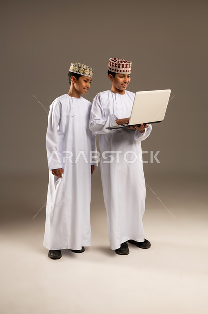 متابعه البرامج الترفيهيه المفضلة بإيماءات السرور والاندماج، بورتريه لولدان توأم عربيان خليجيان عمانيان يرتديان الدشداشة والكمة احدهما يقوم باستخدام جهاز الحاسوب المحمول، الاستفادة من التقنيات الحديثة في تزويد الأجيال بالمعلومات المفيدة، خلفية بيج .