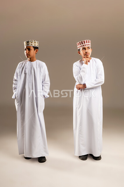 Contemplation and concentration to find a solution to a problem, feeling bored and distressed, problem solving and decision making, portrait of two Arab Gulf Omani twin boys wearing dishdasha and kummah with gestures of thinking, confusion and annoyance, full body image, beige background.