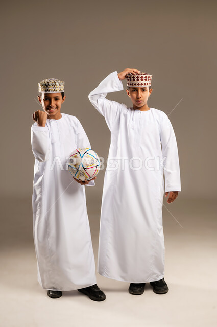 Gestures of loss and defeat, passion for watching sports matches, looking at the camera with interaction, engagement, and enthusiasm, a portrait of two Arab Gulf Omani twin boys wearing a dishdasha and a kummah, a beige background.