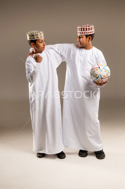 الشغف بمتابعة المباريات الرياضية, النظر إلى الكاميرا بتفاعل واندماج وحماس, السرور والبشاشة لتحقيق الفوز والانتصار, بورتريه لولدان توءم عربيان خليجيان عمانيان يرتديان الدشداشة والكمة , خلفية بيج .