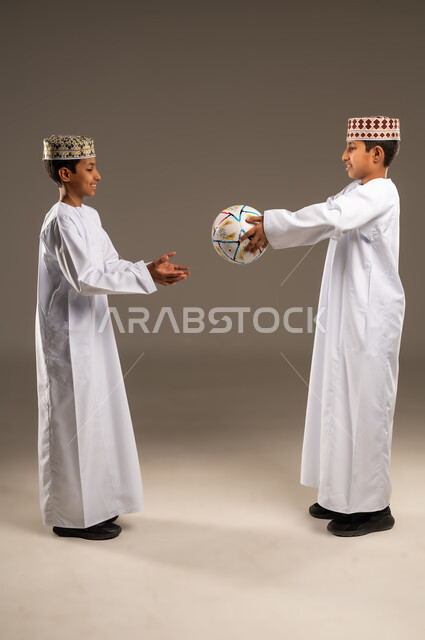 Playing football, gestures of interaction, engagement, and enthusiasm, passion for watching sports matches, side portrait of two Omani Gulf Arab twin boys wearing dishdashas and kummah, beige background.