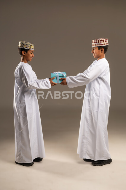 التعبير عن المحبة والألفة بين الأخوان, تقديم الهدايا التذكارية, أعياد ومناسبات ومفاجآت سعيدة, بورتريه من الجانب لولدان توأم عربيان خليجيان عمانيان يرتديان الدشداشة والكمة ، يقوم بتقديم الهدية لأخيه التوأم , صورة الجسم بالطول الكامل, خلفية رمادية .