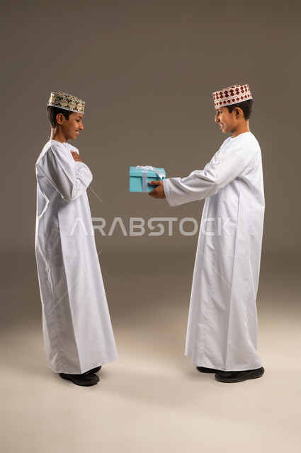 تقديم الهدايا التذكارية, التعبير عن المحبة والألفة بين الأخوان, أعياد ومناسبات ومفاجآت سعيدة, بورتريه من الجانب لولدان توأم عربيان خليجيان عمانيان يرتديان الدشداشة والكمة ، يقوم بتقديم الهدية لأخيه التوأم , صورة الجسم بالطول الكامل, خلفية رمادية .