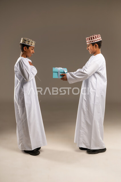 التعبير عن المحبة والألفة بين الأخوان, أعياد ومناسبات ومفاجآت سعيدة, تقديم الهدايا التذكارية, بورتريه من الجانب لولدان توأم عربيان خليجيان عمانيان يرتديان الدشداشة والكمة ، يقوم بتقديم الهدية لأخيه التوأم , صورة الجسم بالطول الكامل, خلفية رمادية .