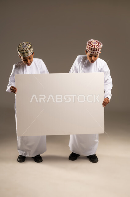لوحة فنية فارغة، موك اب أبيض، مفهوم التوأم الحقيقي المتطابق، استخدام لوحات توضيحية ووسائل تعليمية، بورتريه لولدان توأم عربيان خليجيان عمانيان يرتديان الدشداشة والكمة يحملان موك اب مستطيل وينظران له ، خلفية بيج .