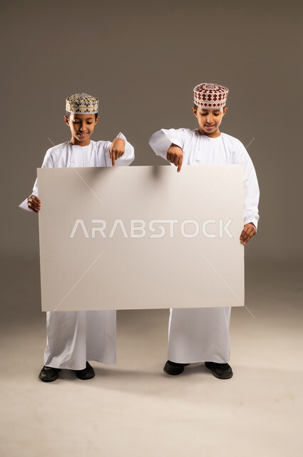 إعلانات وعروض تسويقية, لوح اعلاني فارغ، استخدام لوحات توضيحية ووسائل تعليمية، بورتريه لولدان توأم عربيان خليجيان عمانيان يرتديان الدشداشة والكمة يحملان موك اب مستطيل ويشيران له بإصبع السبابة ، خلفية بيج .