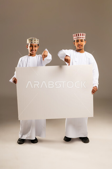 لوح اعلاني فارغ، استخدام لوحات توضيحية ووسائل تعليمية، إعلانات وعروض تسويقية, بورتريه لولدان توأم عربيين خليجيين عمانيان يرتديان الدشداشة والكمة يحملان موك اب مستطيل ويشيران له بإصبع السبابة ، خلفية بيج .