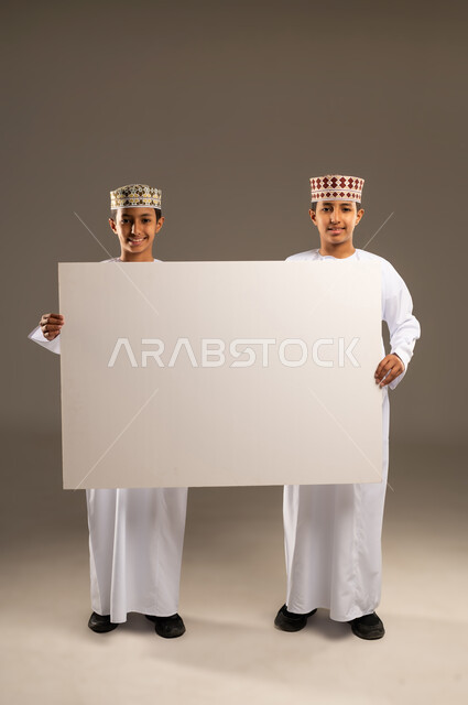 Using illustrations and educational tools, advertisements, and marketing displays, a blank billboard, a portrait of two Omani Gulf Arab twin boys wearing dishdashas and kummahs holding a rectangular mockup, against a beige background.