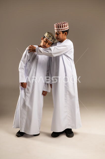 مفهوم التعاطف والمودة بين الأشقاء, الوقوف بجانب بعضهم البعض في الاوقات الصعبة, الشعور بالحزن و الاكتئاب، بورتريه لولدان توأم عربيان خليجيان عمانيان يرتديان الدشداشة والكمة يضع يده على كتف اخيه التوءم لتخفيف الالم والمواساة، خلفية بيج .