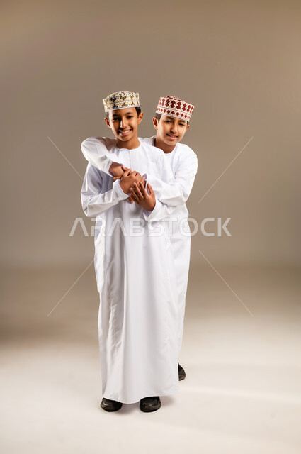 Gestures of love and brotherhood, spending quality time with brothers, elegance and attention to one's appearance, a portrait of two smiling Omani Gulf Arab twin boys wearing the dishdasha and the kummah, standing back to back with gestures of joy, looking at the camera with expressions of happiness and joy, beige background.