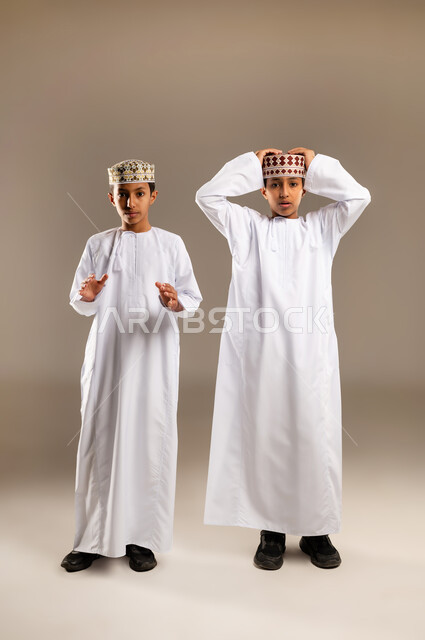 مفهوم التعجب والاندهاش من حدث ما ,النظر الى الكاميرا بايماءات الذعر والخوف ، بورتريه لولدان توأم عربيان خليجيان عمانيان يرتديان الدشداشة والكمة بتعابير الذهول والصدمة ، التفاجؤ من شيء ، الجسم بالطول الكامل، خلفية بيج .