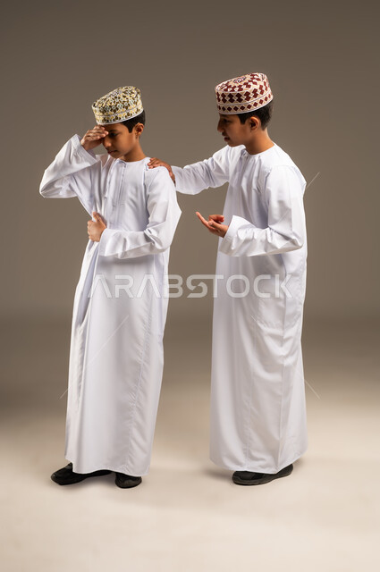Feeling sad and depressed, the concept of empathy and affection between siblings, standing by each other in difficult times, portrait of two Arab Gulf Omani twin boys wearing dishdasha and kummah putting his hand on his twin brother's shoulder to relieve pain and comfort, beige background.