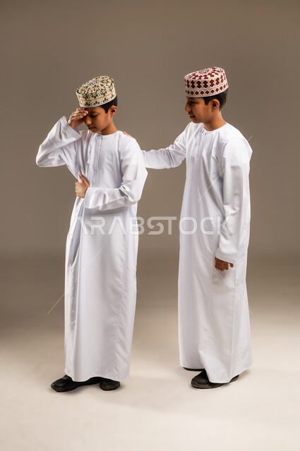 الوقوف بجانب بعضهم البعض في الاوقات الصعبة, الشعور بالحزن و الاكتئاب، مفهوم التعاطف والمودة بين الأشقاء, بورتريه لولدان توأم عربيان خليجيان عمانيان يرتديان الدشداشة والكمة يضع يده على كتف اخيه التوءم لتخفيف الالم والمواساة، خلفية بيج .