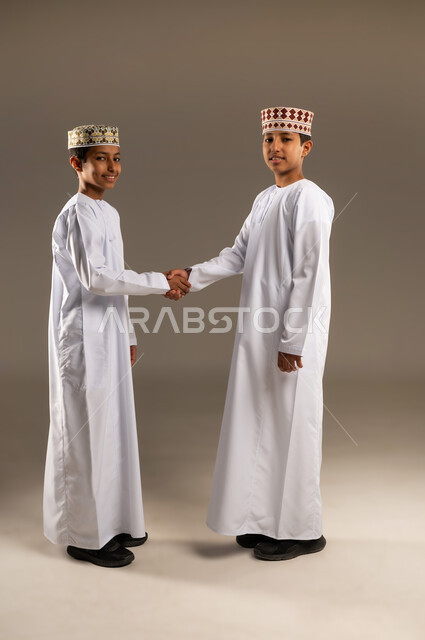 النظر الى الكاميرا بتعابير السرور ، ايماءات وجه ويدين تدل على السعادة، الاتفاق عل شئ ما ، بورتريه لولدان عربيان خليجيان عمانيان يتصافحان بحرارة، العلاقات الاخويه المترابطة، تعارف وصداقات، خلفية بيج .