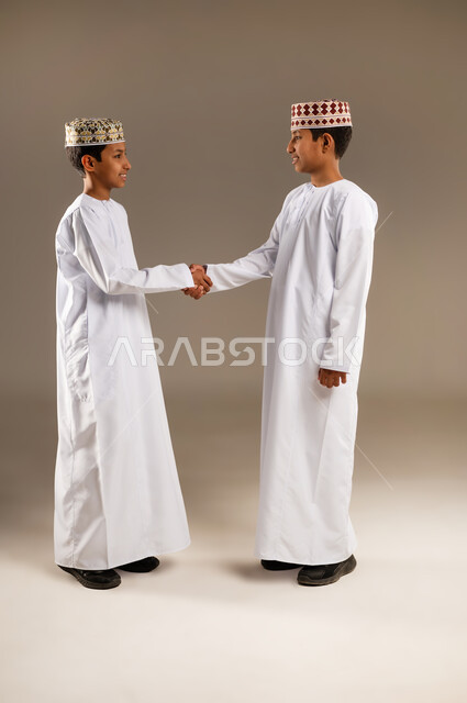 ايماءات وجه ويدين تدل على السعادة، الاتفاق عل شئ ما ، بورتريه لولدان عربيان خليجيان عمانيان يتصافحان بحرارة، العلاقات الاخويه المترابطة، النظر الى الكاميرا بتعابير السرور ، تعارف وصداقات، خلفية بيج .