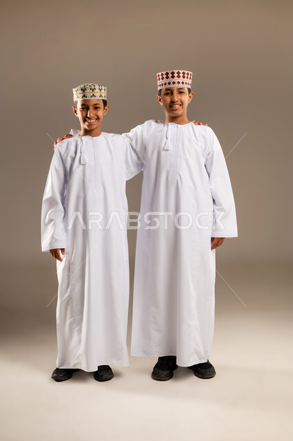 قضاء أوقات ممتعة مع الأخوة، الأناقه والاهتمام بالمظهر الخارجي،  بورتريه  لولدان توأم عربيان خليجيان عمانيان مبتسمان يرتديان الدشداشة والكمة يقفان باستقامة وثقه بالنفس، النظر الى الكاميرا بايماءات السعادة والمرح، خلفية بيج .