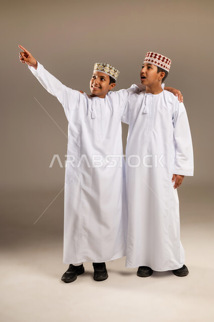 تعابير يدين تشير الى موقع معين, حركات تدل على هذا المكان (هنا), مفهوم لغة الجسد والإشارات، بورتريه لولدان توءم عربيان خليجيان عمانيان مبتسمان يرتديان الدشداشة والكمة ويشيران بأصابع السبابة إلى شيء ما بإيماءات السرور، خلفية بيج.