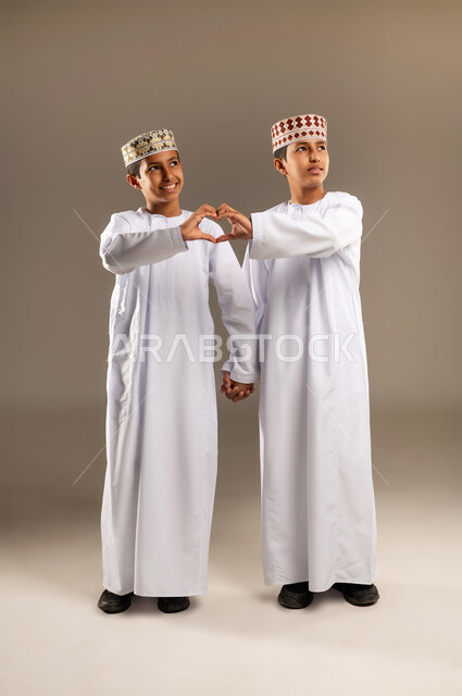إشارات يدين على شكل قلب, المحبة والترابط والألفة, ايماءات تدل على الشعور بالحب، العلاقة الايجابية الاخوية والحب والمودة، بورتريه لولدان توأم عربيان خليجيان سعوديان يرتديان الدشداشة والكمة ينظران الى الاعلى , صورة الجسم بالطول الكامل, خلفية بيج .