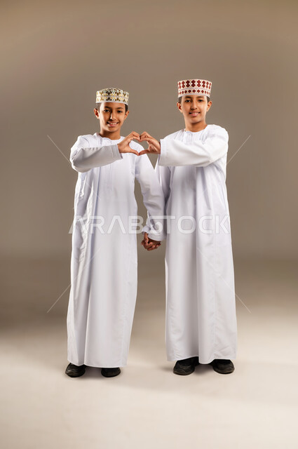 ايماءات تدل على الشعور بالحب، المحبة والترابط والألفة, العلاقة الايجابية الاخوية والحب والمودة، إشارات يدين على شكل قلب, بورتريه لولدان توأم عربيان خليجيان سعوديان يرتديان الدشداشة والكمة, صورة الجسم بالطول الكامل, خلفية بيج .