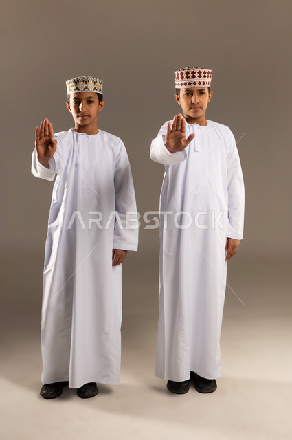 The concept of refusal, abstention and discomfort, a portrait of two Arab Gulf Omani twin boys wearing a dishdasha and a kummah, extending their hands forward with a raised palm, gestures of forbidding something, a request to stop and not approach, expressions of firmness and seriousness, full-length body image, beige background.