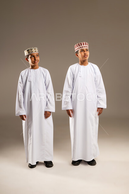 Looking at a specific location above, body language concept, portrait of two smiling Omani Gulf Arab twin boys wearing dishdasha and kummah looking at something with gestures of pleasure, beige background.