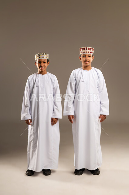 النظر الى الكاميرا بايماءات السعادة والمرح، قضاء أوقات ممتعة مع الأخوة، الأناقه والاهتمام بالمظهر الخارجي، بورتريه  لولدان توأم عربيان خليجيان عمانيان مبتسمان يرتديان الدشداشة والكمة يقفان باستقامة بتعابير الثقة بالنفس، خلفية بيج .