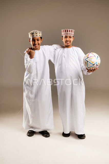 Looking at the camera with interaction, engagement and enthusiasm, passion for following sports matches, joy and happiness for achieving victory and triumph, portrait of two Arab Gulf Omani twin boys wearing the dishdasha and the kummah, beige background.