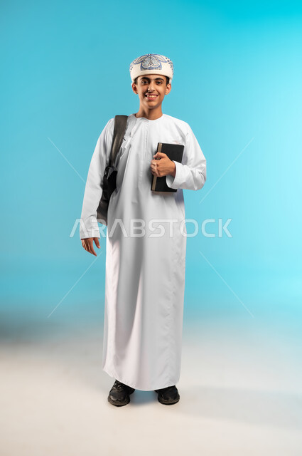 المناهج العلمية التعليمية، مفهوم العودة إلى المدارس، الجد والاجتهاد للوصول إلى النجاح، بورتريه لطالب عربي خليجي عماني يرتدي الدشداشة والكمة يحمل حقيبة الظهر المدرسية يمسك بيده كتاب ، خلفية ملونه .