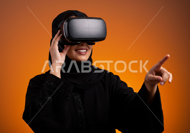 التكنولوجيا الحديثة واستخدام الأجهزة المتطورة، المتعة والتسلية بمحاكاة العالم الخيالي، الاندماج والتفاعل مع الواقع الافتراضي, بورتريه مقرب لامرأة عربية خليجية سعودية محجبة ترتدي نظارة في آر ثلاثية الأبعاد بايماءات التفاعل, خلفية برتقالية