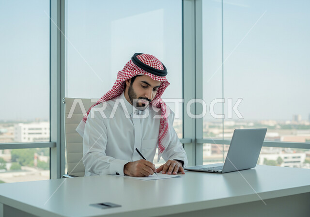 ادارة وانجاز المهام باستخدام جهاز تقني حديث ومتطور, تصوير مقرب لرجل عربي خليجي سعودي مبتسم يرتدي الزي التقليدي يعمل على الحاسوب المحمول, ايماءات وتعابير السرور والسعادة، مهن ووظائف مكتبية سعودية، كتابة وتدوين البيانات والنتائج