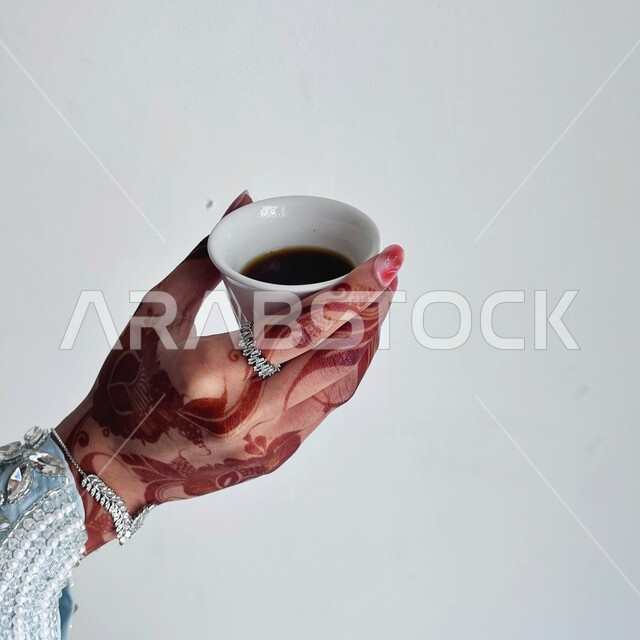 مفهوم الزخرفة والفن اليدوي التقليدي، التجمل والتزين والاهتمام بالمظهر الخارجي، صورة مقربة ليد امرأة خليجية منقوشة بالحناء تحمل بيدها فنجان قهوة، عادات وتقاليد في المناسبات والأفراح السعيدة، نقوش عربيه على اليد، الاستمتاع بالمشروب الساخن