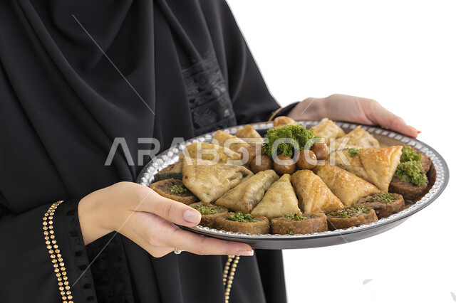 الضيافة التقليدية الشعبية في الأعياد والمناسبات, تقديم حلوى البقلاوة بالفستق, بورتريه مقرب لامرأة عربية خليجية سعودية محجبة مبتسمة ترتدي العباءة السوداء تحمل طبق من الحلويات الشرقية, خلفية بيضاء