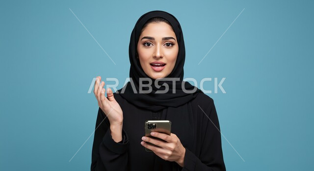 استخدام جهاز تقني حديث, تصفح وسائل التواصل الاجتماعي, بورتريه مقرب لامرأة عربية خليجية سعودية مبتسمة ترتدي الحجاب تمسك بيدها الهاتف المحمول، خلفية ملونة