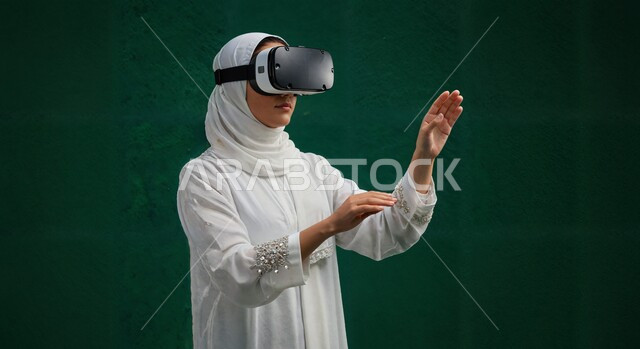 مفهوم التكنولوجيا الحديثة واستخدام الأجهزة المتطورة، المتعة والتسلية بمحاكاة العالم الخيالي، الاندماج والتفاعل مع الواقع الافتراضي, بورتريه مقرب لامرأة عربية خليجية سعودية محجبة ترتدي نظارة في آر ثلاثية الأبعاد بايماءات التفاعل, خلفية خضراء