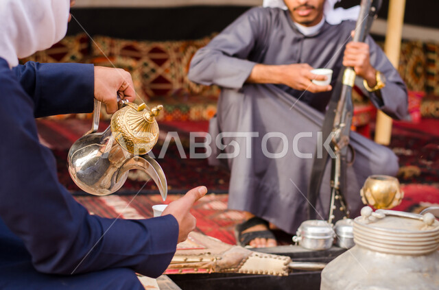 جلسة سعودية تقليدية شعبية، الاستمتاع بشرب القهوة العربية, صورة مقربة ليد رجل عربي سعودي خليجي يقوم بصب القهوة من الدلة لصديقه، قضاء أوقات ممتعة في الأجواء الصحراوية, رحلة تخييم برية وكشتة بر شبابية في النهار