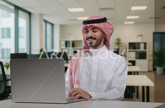 Gestures and expressions of pleasure and happiness, Saudi office professions and jobs, writing and recording data and results, managing and completing tasks using a modern and advanced technical device, a Saudi Arabian Gulf man wearing a traditional shemagh and thobe working on a laptop