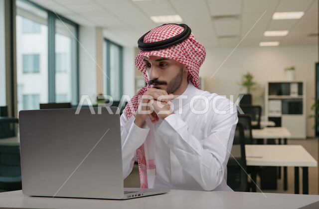ايماءات وتعابير التركيز والاندماج، مهن ووظائف مكتبية سعودية، كتابة وتدوين البيانات والنتائج، ادارة وانجاز المهام باستخدام جهاز تقني حديث ومتطور, رجل عربي خليجي سعودي يرتدي الشماغ والثوب التقليدي يعمل على الحاسوب المحمول يضع يديه تحت ذقنه