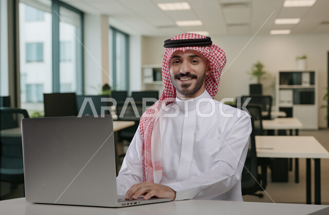 النظر الى الكاميرا بايماءات وتعابير السرور والسعادة، مهن ووظائف مكتبية سعودية، كتابة وتدوين البيانات والنتائج، ادارة وانجاز المهام باستخدام جهاز تقني حديث ومتطور, رجل عربي خليجي سعودي يرتدي الشماغ والثوب التقليدي يعمل على الحاسوب المحمول