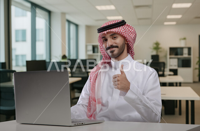 ايماءات وتعابير الاعجاب والرضا والقبول، مهن ووظائف مكتبية سعودية، كتابة وتدوين البيانات والنتائج، ادارة وانجاز المهام باستخدام جهاز تقني حديث ومتطور, رجل عربي خليجي سعودي يرتدي الشماغ والثوب التقليدي يعمل على الحاسوب المحمول يرفع اصبع الابهام للاعلى
