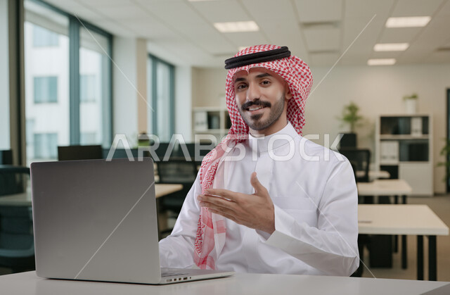 الاشارة بكف اليد، ادارة وانجاز المهام باستخدام جهاز تقني حديث ومتطور, رجل عربي خليجي سعودي يرتدي الشماغ والثوب التقليدي يعمل على الحاسوب المحمول, ايماءات وتعابير السرور والسعادة، مهن ووظائف مكتبية سعودية، كتابة وتدوين البيانات والنتائج