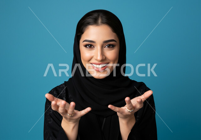 Elegance and interest in outward appearance, the concept of femininity and softness, a close-up portrait of a smiling young Saudi Arabian Gulf woman wearing the hijab, raising her hands up with expressions of enthusiasm and pleasure, looking at the camera with gestures of happiness, blue background