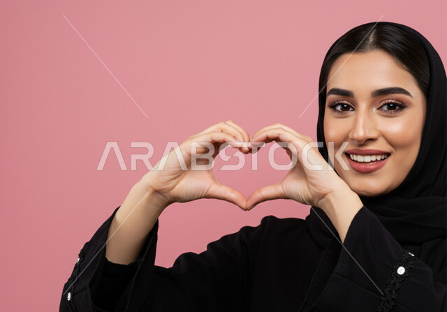 مفهوم المحبة والمودة، اظهار مشاعر الحب والشغف، ايماءات يدين على شكل قلب، بورتريه مقرب لامرأه عربيه خليجيه سعوديه محجبة ترتدي عباءة سوداء تنظر إلى الكاميرا بتعابير السرور والسعادة، خلفية وردية