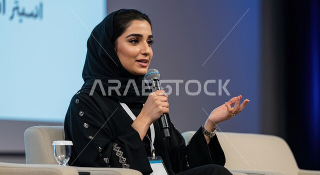اتقان فن الالقاء والخطابة، مهارات التحدث والحوار، إمرأة عربية خليجية سعودية ترتدي الحجاب والعباءة تمسك بيدها مكبر الصوت تشارك في مؤتمر بإيماءات الاندماج والتركيز والثقة بالنفس