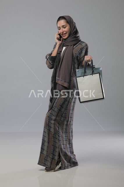اجراء مكالمة هاتفية باستخدام الهاتف المحمول، بورتريه لإمرأة عربية خليجية سعودية ترتدي العباءة والحجاب تحمل بيديها أكياس التسوق، الاستفادة من العروض والتخفيضات الموسميه، تعابير وايماءات السرور والسعادة، الجسم بالطول الكامل، خلفية رمادية