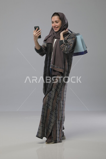 اجراء مكالمة فيديو هاتفية باستخدام الهاتف المحمول، بورتريه لإمرأة عربية خليجية سعودية ترتدي العباءة والحجاب تحمل بيديها أكياس التسوق، الاستفادة من العروض والتخفيضات الموسميه، تعابير وايماءات السرور والسعادة، الجسم بالطول الكامل، خلفية رمادية