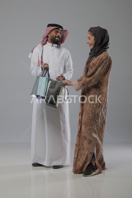 Taking advantage of seasonal offers and discounts, expressions and gestures of pleasure and happiness, buying gifts and supplies, portrait of a Saudi Arabian Gulf couple holding shopping bags in their hands, full body, gray background